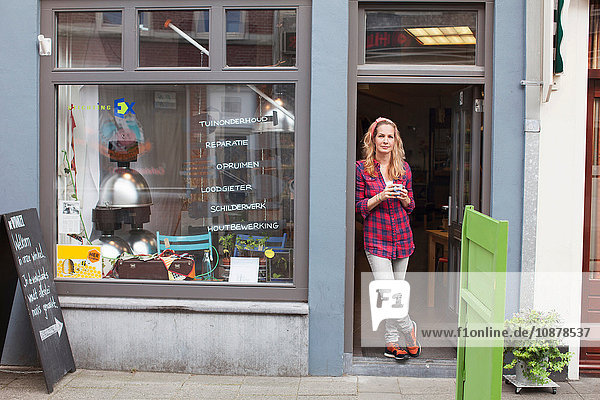 Frau steht in Ladentür und hält Kaffeetasse in der Hand und schaut in die Kamera