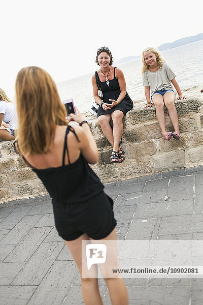 Italien Sardinien Alghero Mädchen (16-17) beim Fotografieren von Mutter und Schwester (14-15) die auf einer Mauer am Meer sitzen.