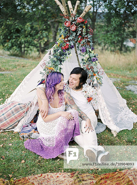 Schweden  Braut und Bräutigam sitzen auf Gras vor weißem Zelt bei Hippie-Hochzeit