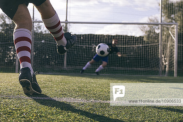 Legs of a footnball player kicking a ball in front of a goal with a goalkeeper