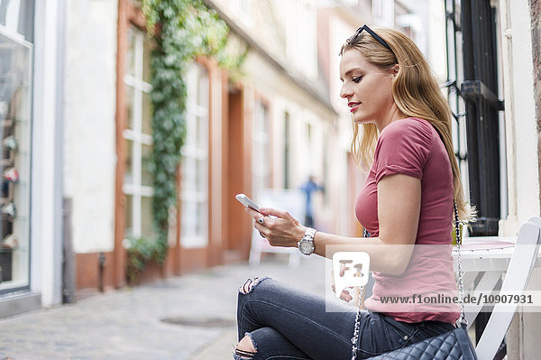 Junge Frau sitzt auf dem Bürgersteig und schaut auf ihr Smartphone.