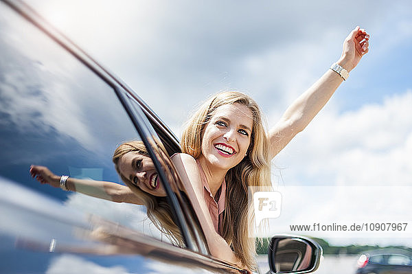 Portrait of smiling young woman leaning out of car window raising arm