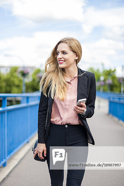 Lächelnde Geschäftsfrau  die auf einer Brücke mit Smartphone läuft