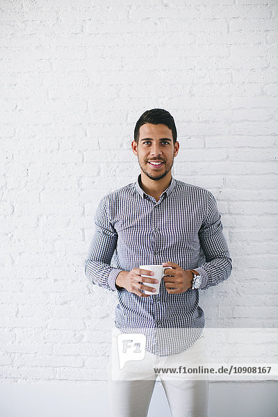 Junger Geschäftsmann steht an der Wand und hält eine Tasse Kaffee hoch.