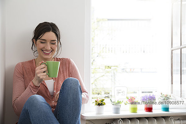 Happy young woman with cup of coffee sitting at the window