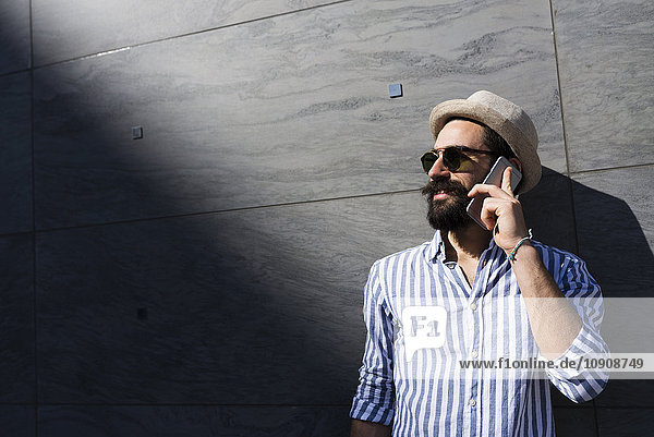 Bartiger junger Mann mit Sonnenbrille und Huttelefonie mit Smartphone