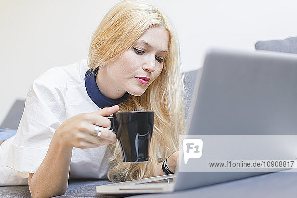 Blond young woman lying on couch with cup of coffee looking at laptop