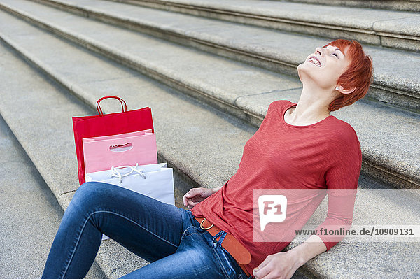 Lächelnde junge Frau entspannt sich auf der Treppe mit Einkaufstaschen neben ihr.