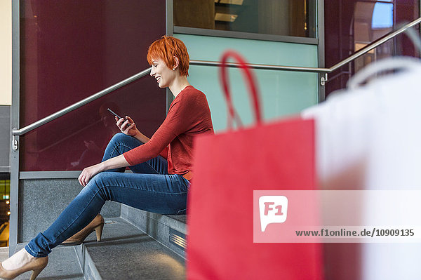 Junge Frau sitzt auf einer Treppe mit Handy und Einkaufstaschen neben ihr.