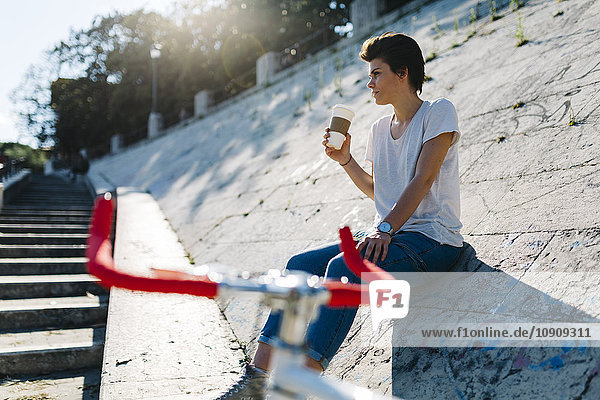 Junge Frau mit Fahrrad und Kaffee zum Sitzen auf geneigter Wand