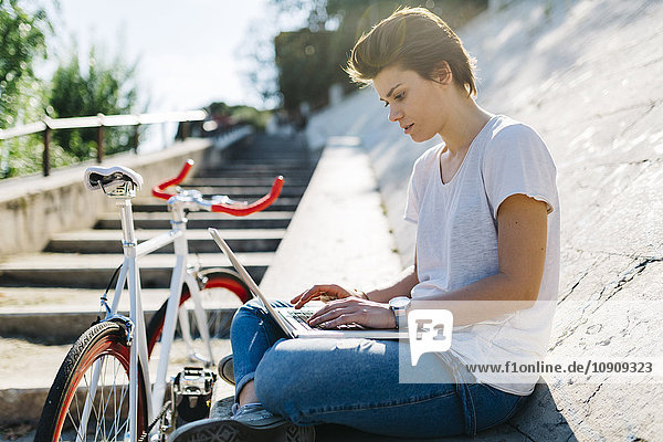 Young woman with bicycle sitting on tilted wall using laptop