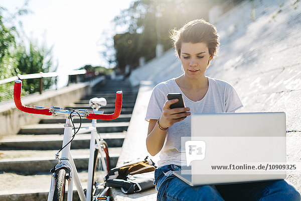 Junge Frau mit Fahrrad im Freien sitzend mit Laptop und Handy