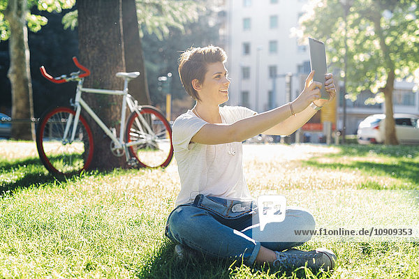 Lächelnde junge Frau sitzt auf der Wiese und nimmt einen Selfie mit digitalem Tablett.
