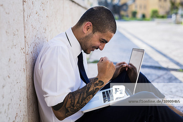 Happy young businessman sitting on the ground looking at his laptop