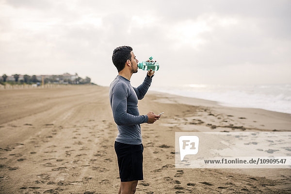 Sportlicher junger Mann beim Trinken aus der Flasche am Strand
