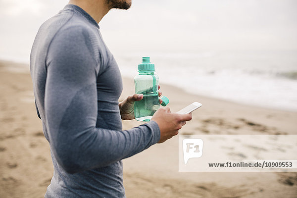 Sportlicher junger Mann mit Handy und Trinkflasche am Strand