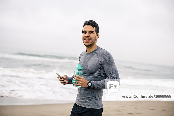 Lächelnder Sportler mit Handy und Trinkflasche am Strand