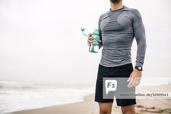 Sportlicher junger Mann mit Trinkflasche am Strand stehend