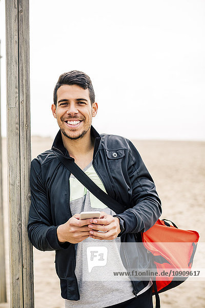 Porträt eines lächelnden jungen Mannes  der sich gegen die Stange am Strand lehnt.
