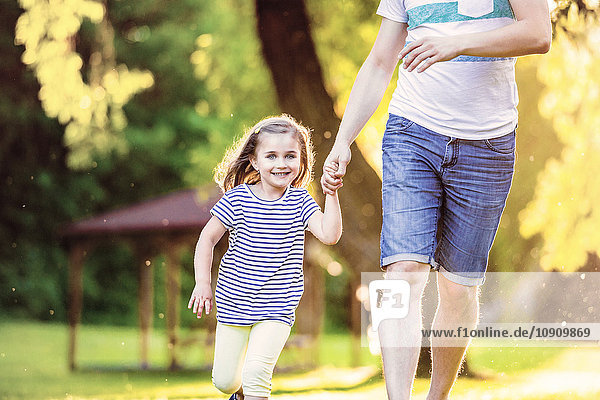 Porträt eines lächelnden kleinen Mädchens  das mit seinem Vater Hand in Hand im Park läuft.