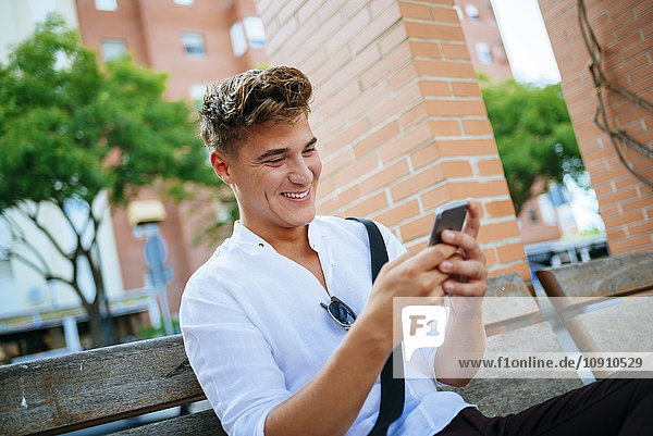 Happy young man sitting on bench looking at smartphone