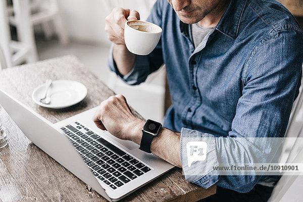 Mature man drinking coffee while working at laptop  looking at smartwatch