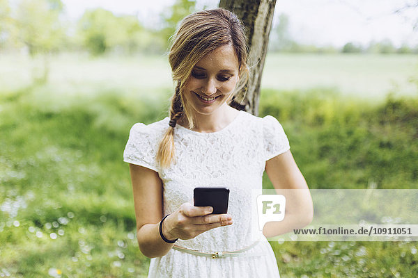 Lächelnde Frau  die sich an den Baumstamm lehnt und ihr Smartphone ansieht