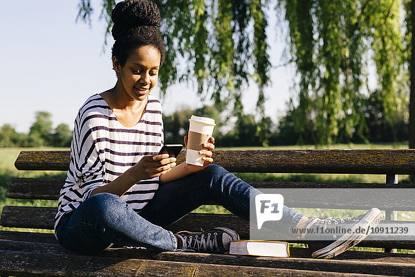 Junge Frau sitzt auf einer Parkbank mit einem Buch und Kaffee  um sich das Smartphone anzusehen.