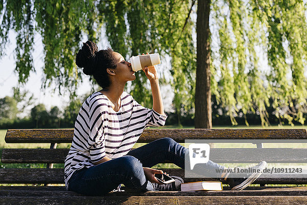 Junge Frau sitzt auf der Parkbank und trinkt Kaffee zum Mitnehmen