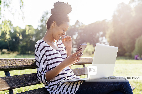 Laughing young woman sitting on park bench with smartphone and laptop