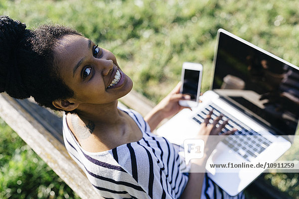 Porträt einer lächelnden Frau auf der Parkbank mit Laptop und Smartphone