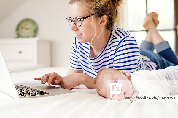 Mother with baby lying on bed working with laptop