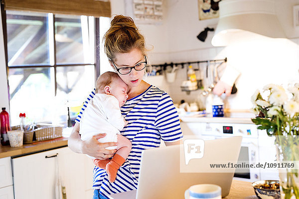 Mutter mit Baby in der Küche mit Blick auf Laptop