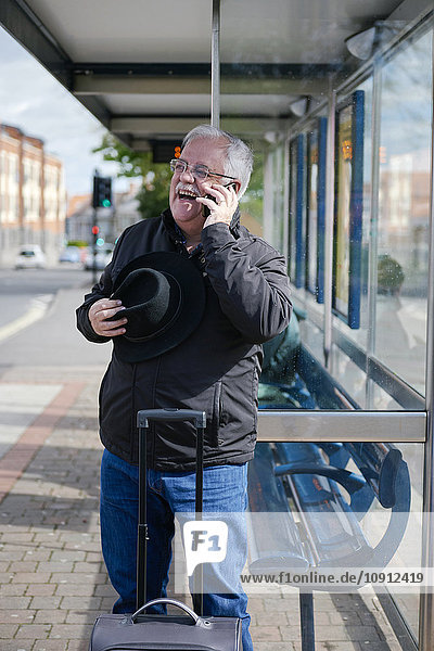 UK  Bristol  Porträt eines lachenden älteren Mannes  der mit dem Smartphone telefoniert  während er an der Bushaltestelle wartet.