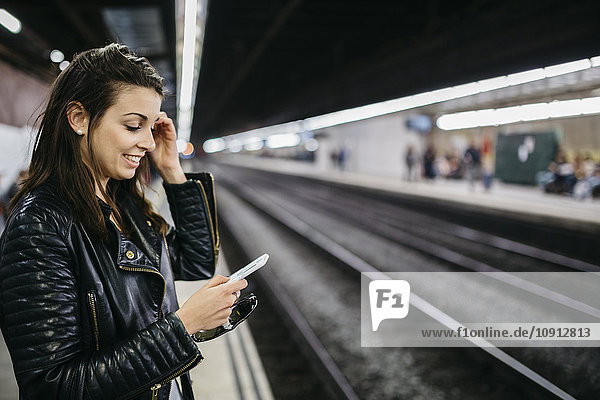 Lächelnde junge Frau auf dem Bahnhof mit Blick aufs Handy