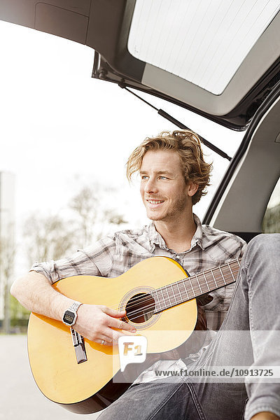 Porträt eines lächelnden jungen Mannes  der auf der Rückseite seines Autos sitzt und Gitarre spielt.