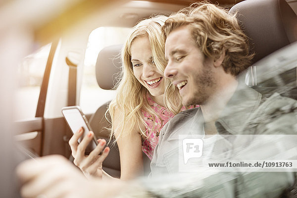 Lachendes junges Paar sitzt in einem Auto und schaut zusammen auf das Smartphone.