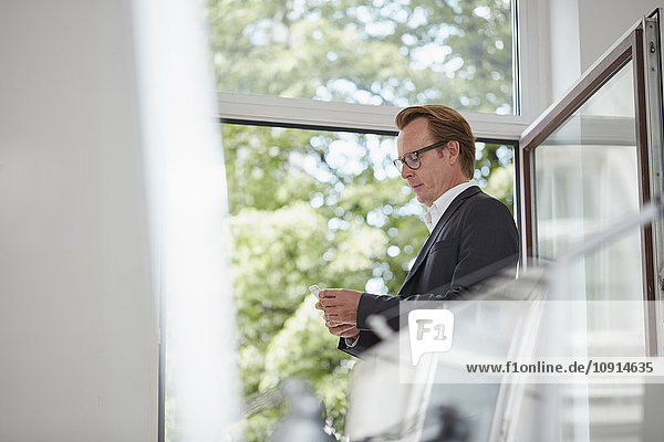 Businessman standing in front of open window looking at his smartphone