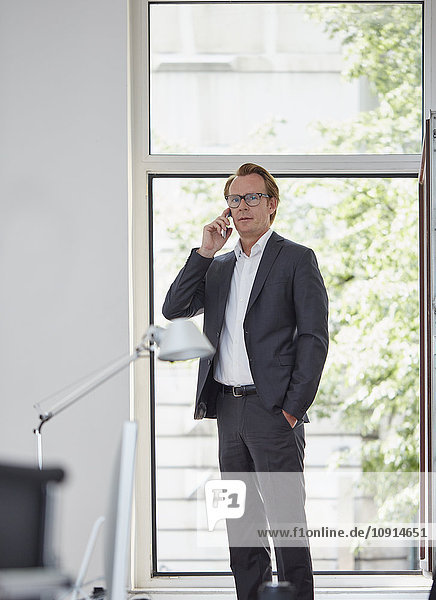 Businessman in his office standing in front of open window telephoning with smartphone