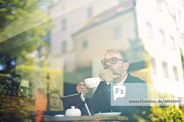 Nachdenklicher Geschäftsmann mit einer Tasse Kaffee in einem Café hinter der Fensterscheibe