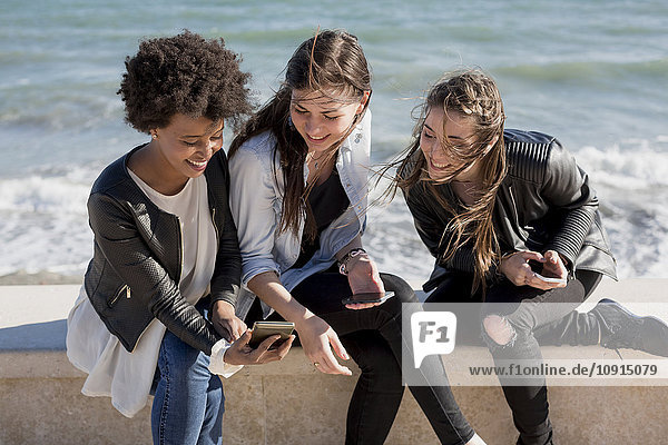 Drei junge Frauen sitzen an der Wand und schauen auf das Smartphone.