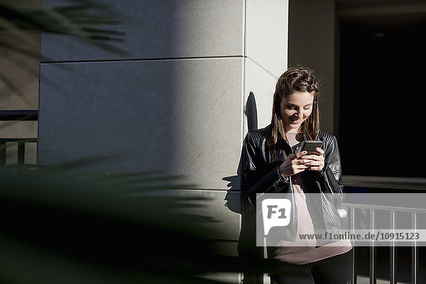 Junge Frau  die sich mit dem Smartphone an die Wand lehnt