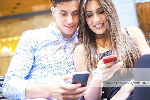 Porträt eines jungen Paares  das auf einer Bank sitzt und sein Smartphone betrachtet.