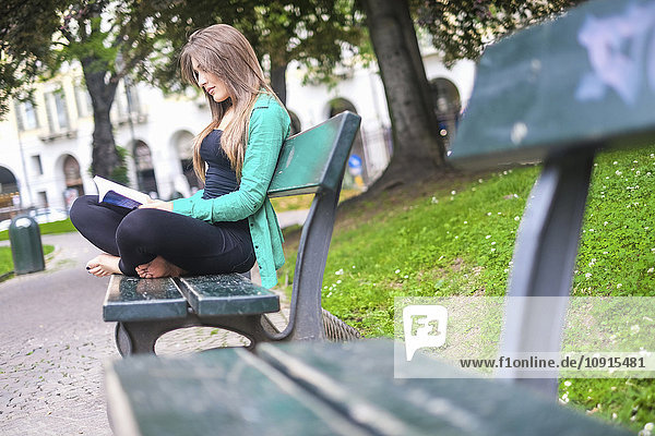 Junge Frau beim Lesen eines Buches  sitzt auf einer Bank im Park
