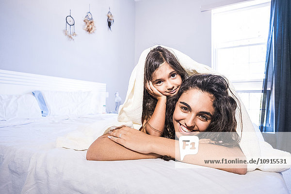 Porträt eines Teenagermädchens und ihrer kleinen Schwester  die unter einer Decke auf dem Bett liegen.