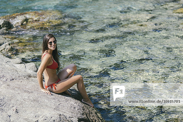 Frau im roten Bikini auf Stein sitzend  lächelnd