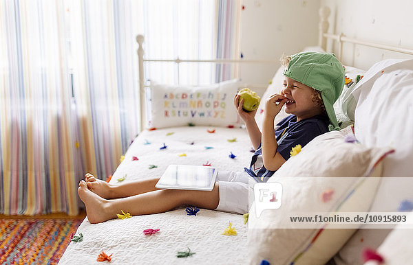 Lachender kleiner Junge auf der Couch sitzend mit Apfel und digitalem Tablett