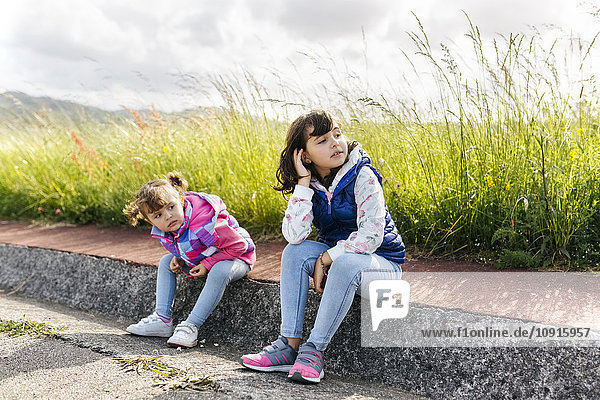 Zwei kleine Schwestern sitzen auf dem Bordstein und schauen in die Ferne.