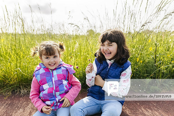 Zwei lachende kleine Schwestern sitzen vor einer Wiese.
