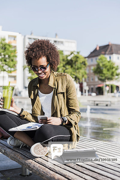 Junge Frau sitzt auf einer Bank mit Getränk und Smartphone und liest Notizen.